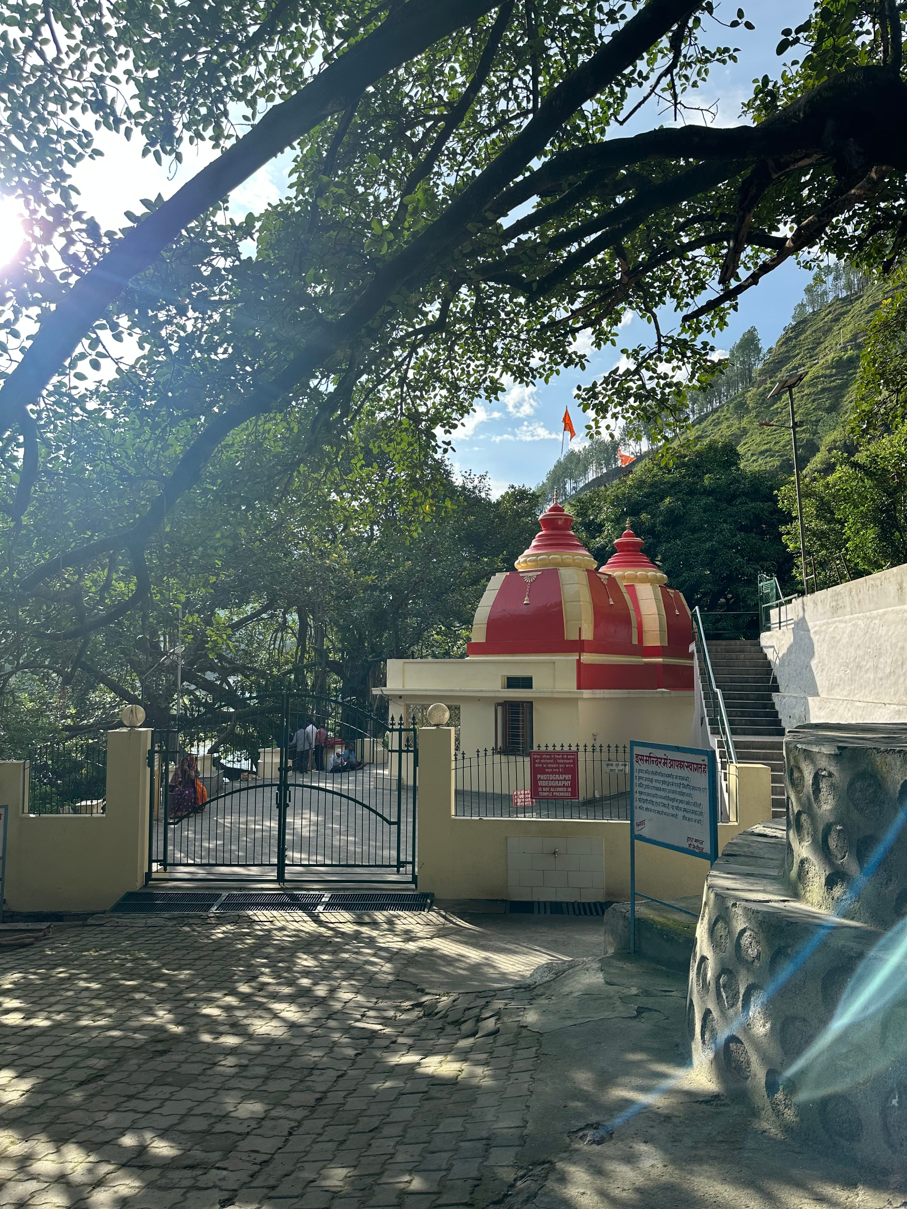 A small red and cream temple beneath trees on a hillside road